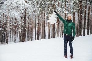 beau modèle masculin d'âge moyen porte un bonnet de fourrure chaud avec des oreillettes et un manteau rembourré en duvet vert, tient un sapin artificiel blanc, se tient sur un sol recouvert de neige, a une expression heureuse photo