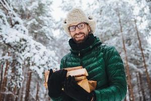 beau mâle barbu en lunettes et chapeau chaud avec anorak, tient du bois de chauffage, pose contre des arbres recouverts de neige blanche étincelante, passe du temps en forêt. tir extérieur d'un homme heureux repose dans la forêt photo