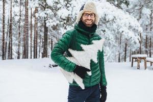 un homme barbu à l'air agréable porte des vêtements chauds, tient un sapin blanc, se dresse sur fond de forêt d'hiver, se promène avec des amis par temps glacial, étant de bonne humeur. les gens et la nature photo