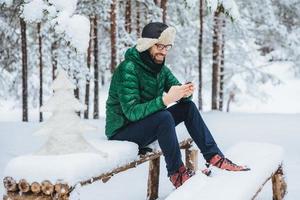 un homme joyeux aime la communication sur un smartphone et la solitude, une atmosphère calme, est assis sur un banc en bois recouvert de neige dans la forêt d'hiver, heureux de recevoir un message d'un ami proche photo