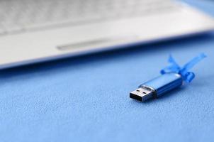 carte mémoire flash usb bleu brillant avec un arc bleu se trouve sur une couverture de tissu polaire bleu clair doux et poilu à côté d'un ordinateur portable blanc. conception de cadeau féminin classique pour une carte mémoire photo