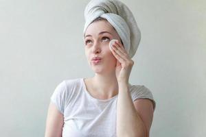 femme souriante dans une serviette sur la tête avec une peau douce et saine enlevant le maquillage avec un tampon de coton isolé sur fond blanc photo