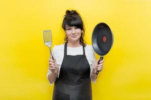 belle femme âgée cuisiner dans un tablier sourit et tient une poêle à frire dans une main dans l'autre spatule, sur fond jaune photo