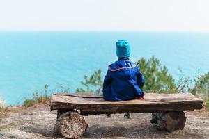 un enfant solitaire se repose sur un banc dans la forêt avec vue sur la mer. photo