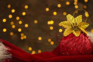 boule de noël rouge avec fleur d'or et chapeau de père noël rouge avec lumières du nouvel an à l'arrière. arrière-plan flou. carte postale de vacances photo
