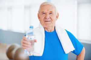 rester en bonne santé heureux homme âgé avec une serviette sur les épaules en tendant une bouteille d'eau tout en se tenant dans un club de santé photo