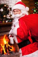 père Noël. père noël traditionnel portant un sac avec des cadeaux et souriant avec un sapin de noël et une cheminée en arrière-plan photo