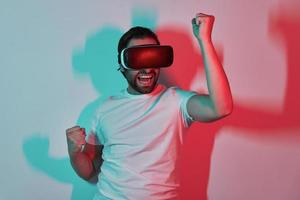 heureux jeune homme dans des lunettes de réalité virtuelle gesticulant avec des ombres colorées en arrière-plan photo