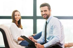 toujours prêt à vous aider. jeune homme confiant en chemise et cravate tenant le presse-papiers et souriant tandis que la femme assise en arrière-plan photo