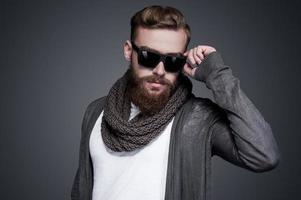 look cool et tendance. beau jeune homme barbu ajustant ses lunettes de soleil et regardant la caméra en se tenant debout sur fond gris photo