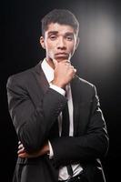 rester calme. jeune homme afro-américain à la mode tenant la main sur le menton en se tenant debout sur fond noir photo
