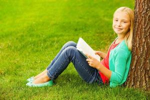 petit rat de bibliothèque. jolie petite fille aux cheveux blonds tenant un livre et souriant assis sur l'herbe verte et se penchant à l'arbre photo