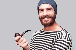 en style marin. beau jeune homme barbu en vêtements rayés tenant une pipe et regardant la caméra avec le sourire en se tenant debout sur fond gris photo