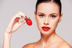 douce tentation. belle jeune femme torse nu tenant une fraise à la main en se tenant debout sur fond gris photo