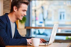 surfer sur le net au café. vue latérale d'un beau jeune homme travaillant sur un ordinateur portable et souriant tout en dégustant un café au café