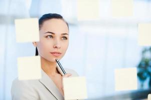 planification de la stratégie commerciale. jeune femme d'affaires réfléchie touchant son menton avec un stylo et regardant les notes adhésives attachées au verre photo