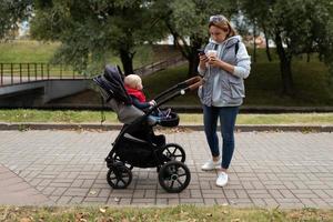une jeune mère boit du café et regarde un téléphone portable tout en marchant avec un bébé dans une poussette autour de la ville photo