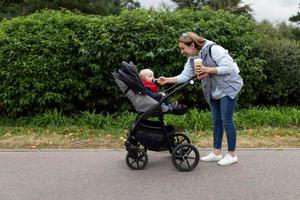 une jeune mère avec une tasse de café dans ses mains avec un sourire sur son visage redresse le mamelon en marchant dans le parc à son petit bébé dans une poussette photo