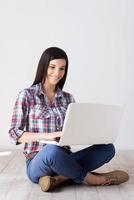 beauté avec ordinateur portable. jolie jeune femme travaillant sur un ordinateur portable et souriant assis sur le plancher de bois franc photo