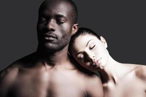 instants sensuels. portrait d'un homme africain torse nu et d'une femme caucasienne se liant l'un à l'autre et gardant les yeux fermés en se tenant debout sur fond gris photo