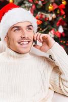 félicitations beau jeune homme en bonnet de noel parlant sur téléphone portable et souriant avec l'arbre de noël en arrière-plan photo