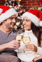 bonne année heureux jeune couple d'amoureux en chapeaux de père noël se liant les uns aux autres et tenant des verres à vin avec l'arbre de noël en arrière-plan photo
