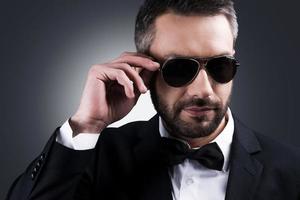 briseur de cœur confiant. portrait d'un bel homme mûr en tenues de soirée ajustant ses lunettes de soleil en se tenant debout sur fond gris photo