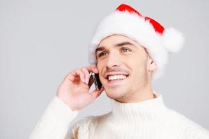 joyeux noël beau jeune homme en bonnet de noel parlant au téléphone mobile et souriant en se tenant debout sur fond gris photo