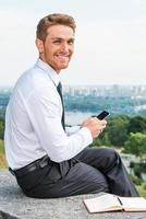 rester connecté à tout moment et n'importe où. jeune homme confiant en chemise et cravate tenant un téléphone portable et souriant tout en étant assis à l'extérieur avec le paysage urbain en arrière-plan photo