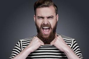 émotions libérées. furieux jeune homme barbu en vêtements rayés étirant son col et criant en se tenant debout sur fond gris photo