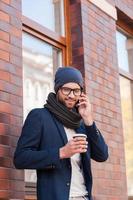 mode de vie de la ville. joyeux jeune homme en tenue décontractée intelligente tenant une tasse de café et parlant au téléphone portable tout en se tenant dans la rue photo