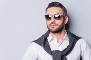 confiant beau. portrait d'un beau jeune homme à lunettes de soleil et vêtements décontractés intelligents en détournant les yeux en se tenant debout sur fond gris photo