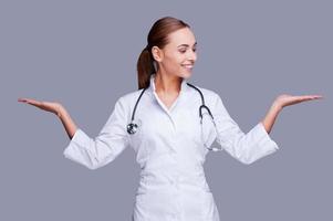 satisfaite de son choix. femme médecin confiante en uniforme blanc tenant des espaces de copie dans ses mains et souriant debout sur fond gris photo