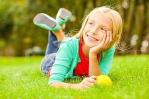 petit rêveur. jolie petite fille se penchant la tête sur la main et regardant loin avec le sourire en position allongée sur l'herbe verte photo