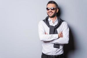 joyeux beau. beau jeune homme à lunettes de soleil et vêtements décontractés intelligents en gardant les bras croisés et souriant en se tenant debout sur fond gris photo