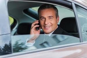 voyager avec confort. heureux homme d'affaires mature parlant au téléphone mobile et souriant assis sur le siège arrière d'une voiture photo