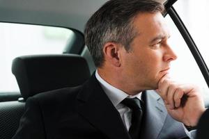 perdu dans ses pensées professionnelles. homme d'affaires mûr et réfléchi tenant la main sur le menton et regardant loin tout en étant assis sur le siège arrière d'une voiture photo