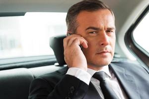 homme d'affaires prospère en voiture. homme d'affaires mature confiant parlant au téléphone mobile et regardant ailleurs assis sur le siège arrière d'une voiture photo