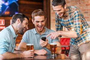 amis s'amusant. trois jeunes hommes heureux en tenue décontractée buvant de la bière dans un pub tandis que l'un d'eux pointe un smartphone et sourit photo