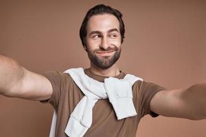 jeune homme espiègle faisant selfie et grimaçant en se tenant debout sur fond marron photo