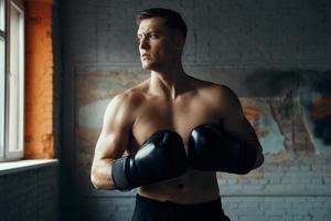 jeune homme confiant dans des gants de boxe regardant loin tout en se tenant dans la salle de sport photo