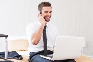 rester en contact avec ses collègues. beau jeune homme en chemise et cravate travaillant sur un ordinateur portable et parlant au téléphone portable assis sur le lit dans la chambre d'hôtel photo