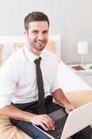 rester connecté à tout moment. beau jeune homme en chemise et cravate travaillant sur un ordinateur portable et souriant assis sur le lit dans la chambre d'hôtel photo