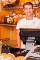 caissier confiant. beau jeune homme caissier en tablier debout près de la caisse enregistreuse et souriant tout en se tenant dans la boulangerie photo