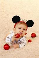 adorable petite fille aux oreilles de souris est allongée sur un plaid beige et joue avec des boules de noël brillantes rouges. photo
