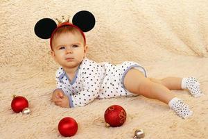 jolie petite fille avec des oreilles de souris sur un plaid beige avec des boules de noël rouges et argentées. photo