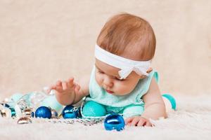 jolie petite fille en robe bleue et blanche joue avec des décorations de noël sur un plaid en peluche beige avec des lumières de noël. photo