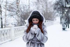 belle jeune femme aux cheveux rouges en manteau de fausse fourrure grise avec capuche sur fond de parc enneigé d'hiver. photo