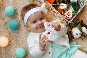 belle petite fille souriante en robe bleue et blanche joue avec un jouet en bois sur un plaid beige avec des décorations et des lumières de noël, vue de dessus. photo