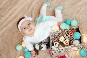 adorable petite fille en robe bleue et blanche est allongée sur un plaid beige, jouant avec des décorations de noël et regardant la caméra. photo